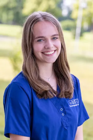 Frau mit langen, dunkelblonden Haaren trägt einen blauenArztkittel. Sie steht im Freien, mit unscharfem Hintergrund aus Bäumen und Wiese. Sie lächelt.