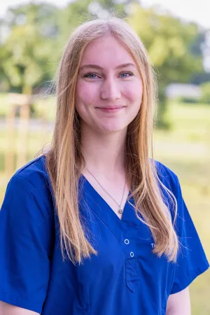 Leicht lächelnde Frau mit langen, blonden Haaren trägt einen blauen Arztkittel. Sie steht im Freien, umgeben von grüner Landschaft.