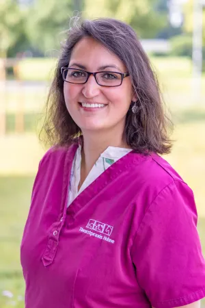 Tierärztin mit schulterlangen, braunen Haaren in einem magenta Kittel. Sie steht im Freien, im Hintergrund sind unscharfe grüne Bäume zu sehen.