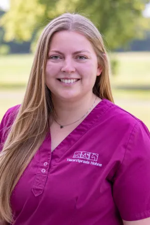 Frau mit langen, blonden Haaren trägt ein lila Oberteil mit Knopfleiste und dem Text 'Tierarztpraxis Hohne' auf der linken Brustseite.