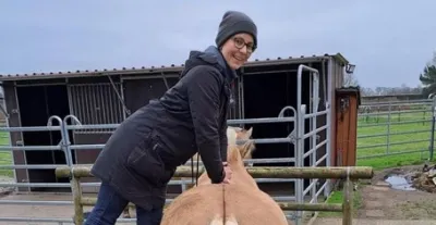 Eine Person steht erhöht und behandelt ein Pferd am Rücken. Die Person trägt eine graue Mütze und einen schwarzen Mantel. Im Hintergrund ist ein Stall sichtbar.