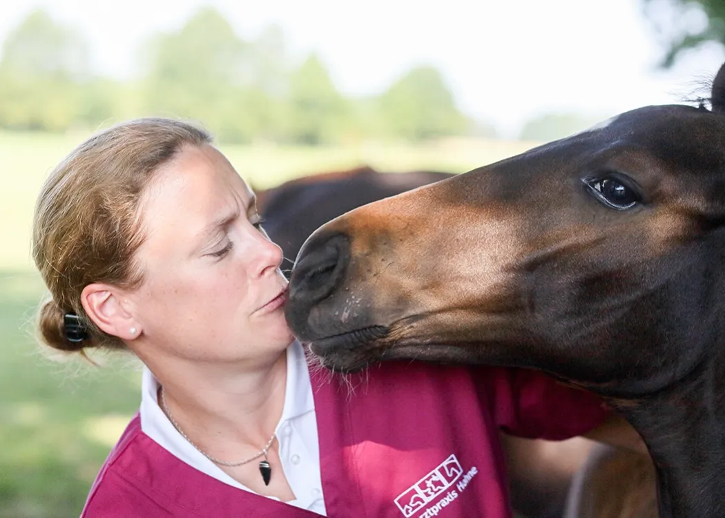 Tierärztin im magenta Oberteil steht im Freien und wird von einem großen braunen Hund neugierig angeschnüffelt und am Gesicht berührt, während sie lächelt – Alltag mit tierischen Patienten in der Tierarztpraxis.