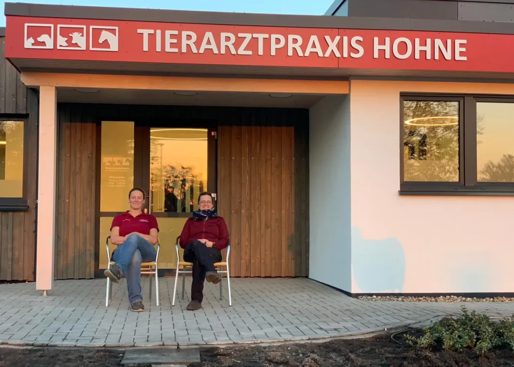 Eingang der Tierarztpraxis Hohne im Harzklint mit rotem Vordach und großem Schild „TIERARZTPRAXIS HOHNE“. Zwei Personen sitzen auf Stühlen vor der Glasfront und lächeln, daneben ein bepflanzter Bereich und helle Fassade.