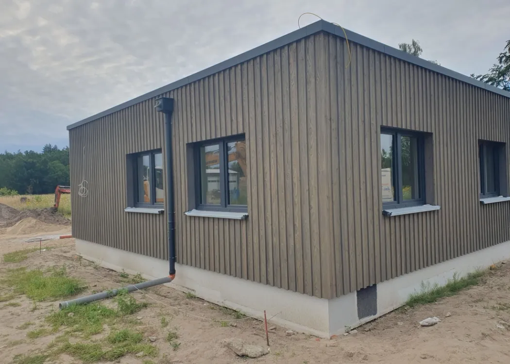 Seitenansicht der Tierarztpraxis Hohne im Harzklint. Fertiggestelltes Gebäude in moderner Holzoptik mit großen rechteckigen Fenstern, umgeben von einer leichten Böschung und sandigem Untergrund.