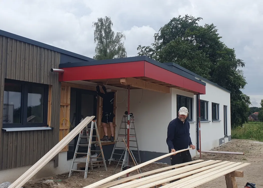 Bauarbeiten am Eingangsbereich der Tierarztpraxis Hohne im Harzklint. Arbeiter montieren ein rotes Vordach, Holzlatten und Baumaterial liegen im Vordergrund, die Fassade ist bereits mit Holz verkleidet.