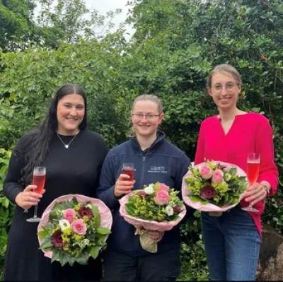 Drei Personen stehen im Freien und halten Blumensträuße in der Hand. Die linke Person trägt ein schwarzes Kleid, die mittlere Person eine dunkelblaue Fleecejacke, die rechte Person ein rotes Oberteil. Alle halten ein Sektglas in der Hand und lächeln.