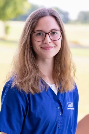 Frau mit langen, lockigen Haaren trägt ein blaues Oberteil mit dem Logo der Tierarztpraxis Hohne. Sie steht im Freien.