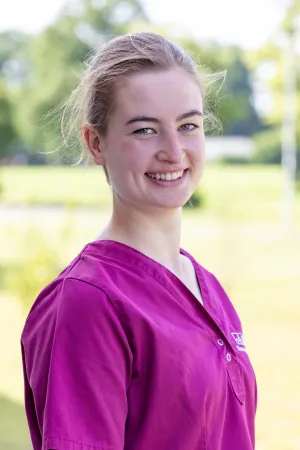 Frau in lila Tierarztpraxis-Kleidung, steht im Freien. Sie hat blonde Haare und trägt ein kurzärmliges Oberteil mit Knöpfen.