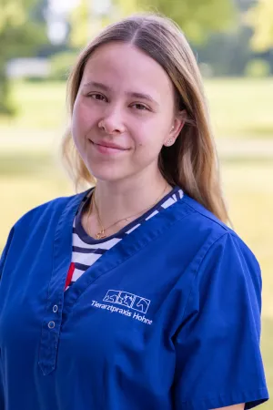 Frau mit blonden Haaren trägt ein blauen Arztkittel und ein gestreiftes Oberteil darunter. Sichtbar ist ein goldener Anhänger um den Hals. Sie steht im Freien und lächelt leicht.