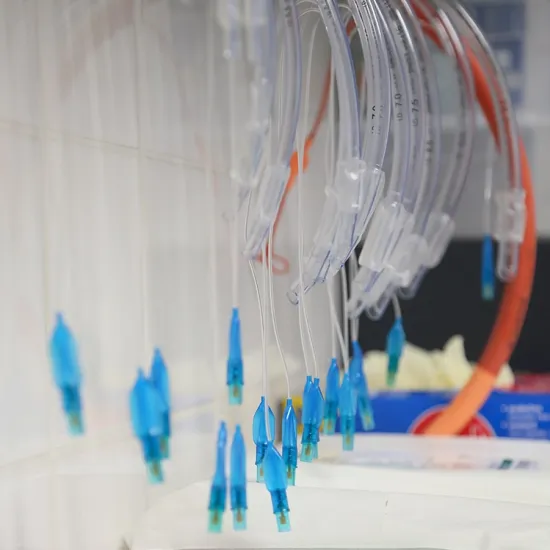 Mehrere transparente medizinische Schläuche mit blauen Anschlüssen hängen an einer Wand. Im Hintergrund sind weitere medizinische Utensilien sichtbar.