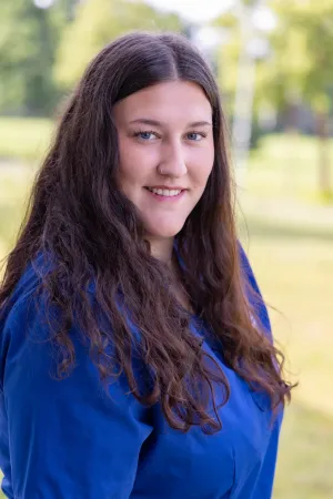 Lächelnde Frau mit dunklen, langen, lockigen Haaren trägt einen blauen Kittel. Sie steht im Freien, mit unscharfem Hintergrund.