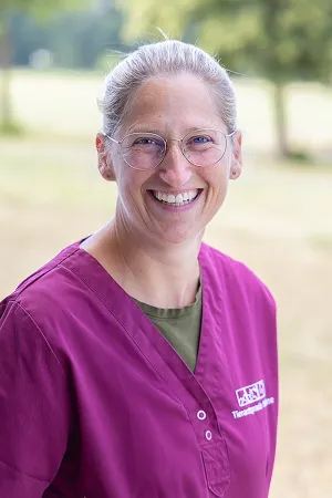 Lächelnde Tierärztin in magenta Kittel, mit blonden, zusammengebundenen Haaren, steht im Freien. Sie trägt eine Brille. Der Hintergrund zeigt unscharfe grüne Bäume.