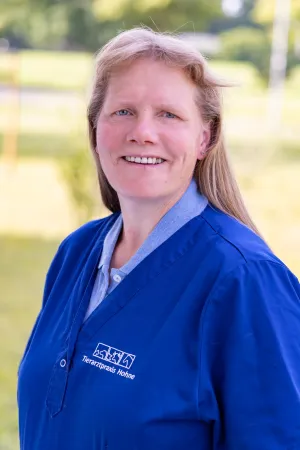 Frau mit langen, blonden Haaren trägt einen blauen Kittel mit dem Logo 'Tierarztpraxis Hohne'. Sie steht im Freien.