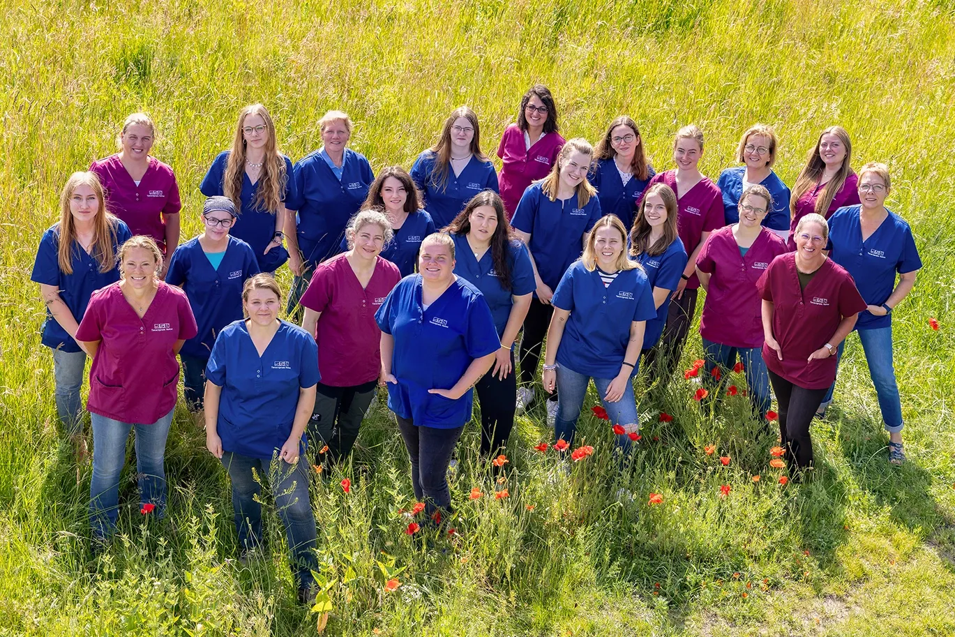 Gruppe von 19 Personen in blauen und magenta Arbeitskleidung steht auf einer Wiese mit roten Blumen
