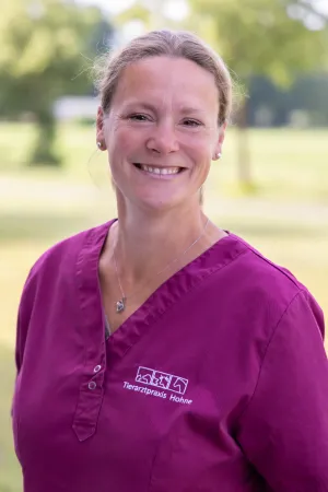 Lächelnde Frau mit dunkelblonden Haaren in magenta Arztkittel mit gesticktem Logo 'Tierarztpraxis Hohne'. Sie steht im Freien, der Hintergrund zeigt unscharfe grüne Bäume.