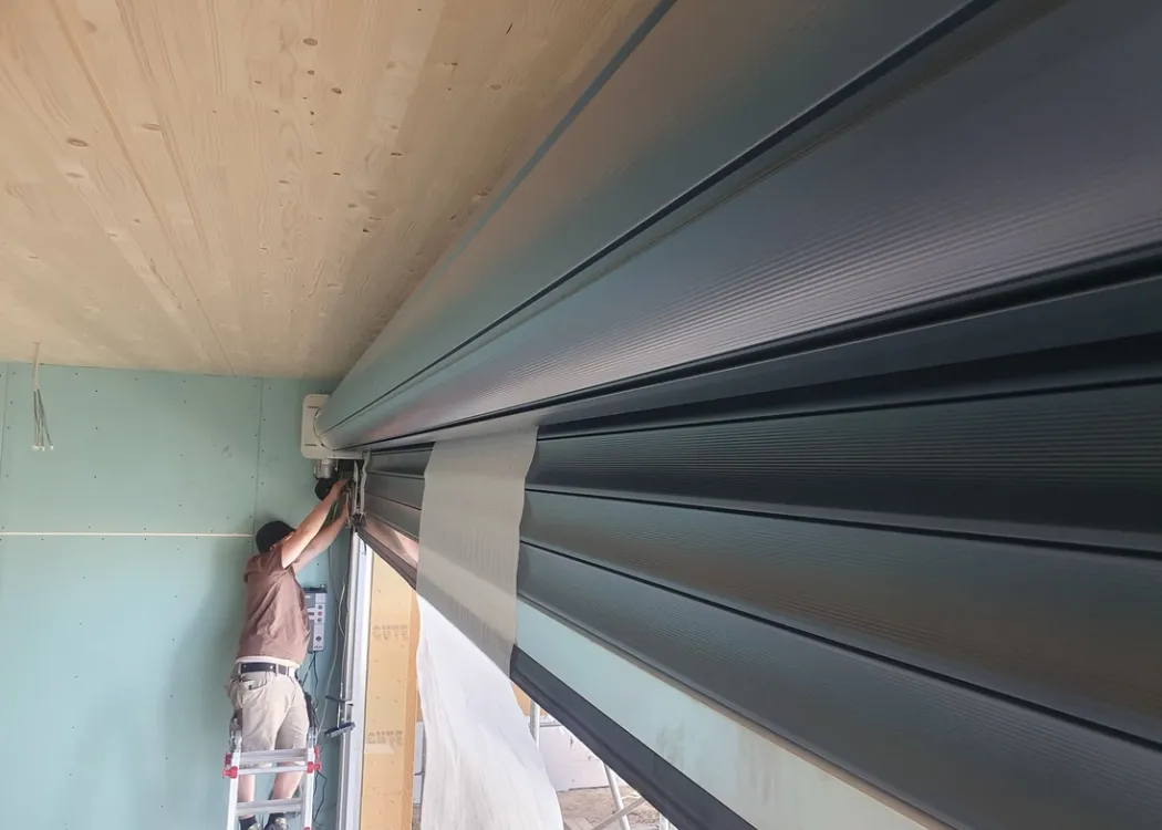 Detailaufnahme einer eingebauten Rollladenkonstruktion in der Tierarztpraxis Hohne im Harzklint. Nahaufnahme von grauen Lamellen unter einer Holzdecke, seitlich sichtbare Baufolie und Rohbaucharakter.