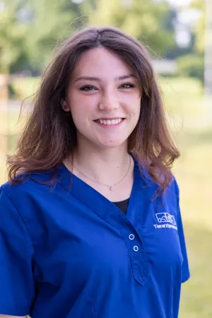 Lächelnde Frau mit langen, braunen Haaren trägt einen blauen Arztkittel mit einem weißen Logo der Tierarztpraxis Hohne auf der linken Brustseite.
