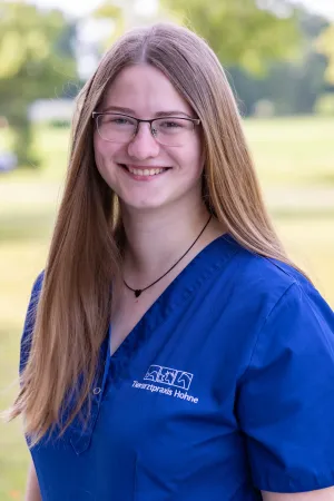 Frau mit langen, blonden Haaren trägt ein blaues Oberteil mit dem Logo 'Tierarztpraxis Hohne'. Hintergrund zeigt unscharfe grüne Bäume.
