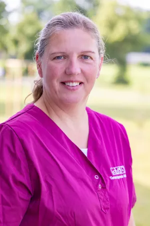 Lächelnde Tierärztin in magenta Kittel, mit langen, hellen, gräulichen Haaren, steht im Freien. Der Hintergrund zeigt unscharfe grüne Bäume.