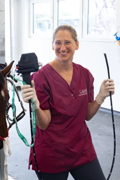 Tierärztin in einem burgunderfarbenen Kittel hält ein schwarzes Bronchoskop in derau linken Hand und einen langen, flexiblen Schlch in der rechten Hand. Die Frau lächelt in die Kamera.