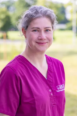 Tierärztin Dr. Katja Eckermann-Mosch trägt ein magenta Oberteil mit Knopfleiste. Sie steht im Freien, umgeben von unscharfem Hintergrund.