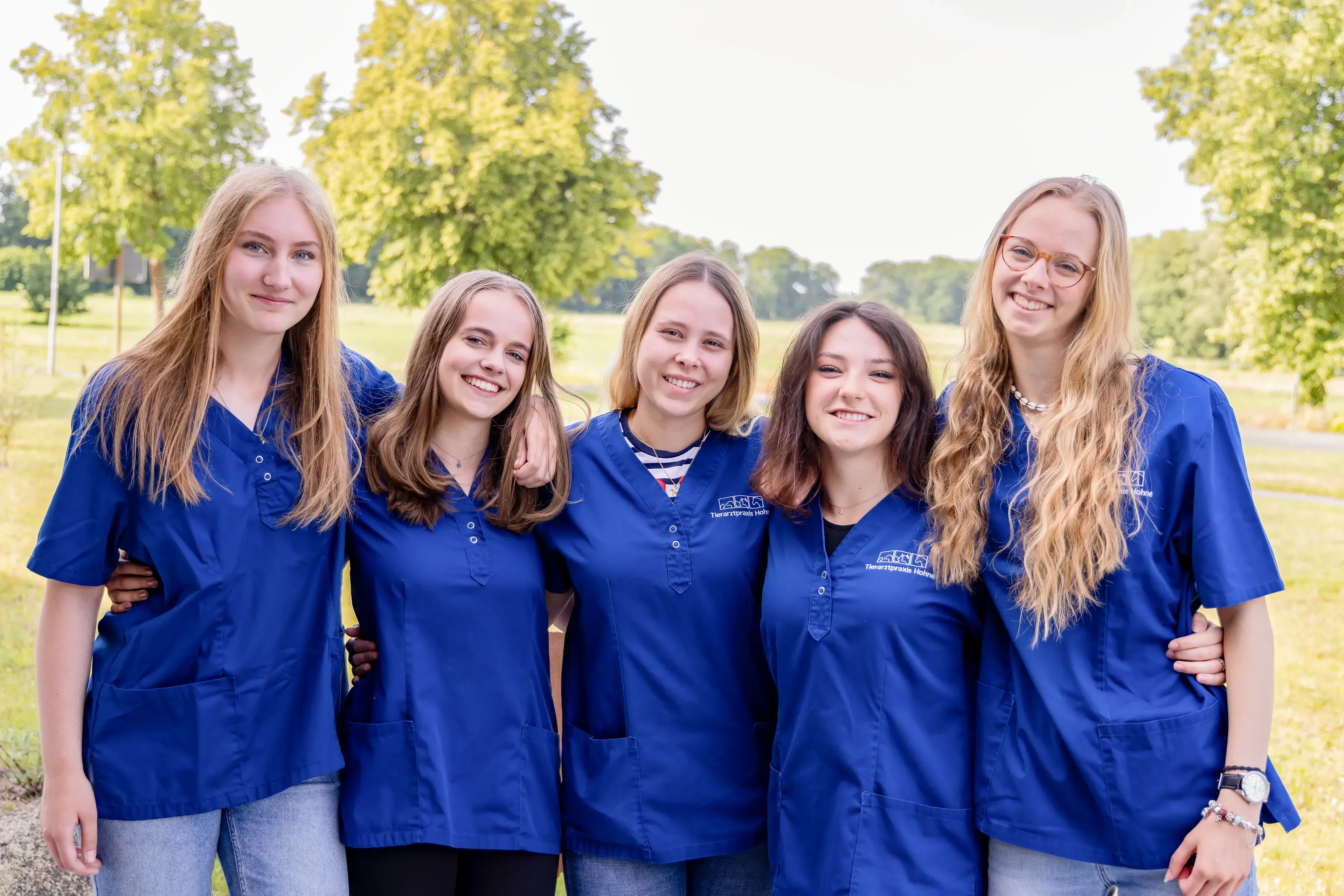 Fünf Personen in blauen Arztkitteln stehen nebeneinander Arm in Arm im Freien. Sie lächeln freudig in die Kamera..