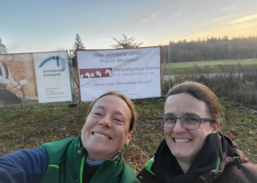Zwei lächelnde Frauen stehen vor einem Baugrundstück mit einem großen Werbebanner. Sie tragen Outdoor-Kleidung, die Stimmung wirkt freudig.