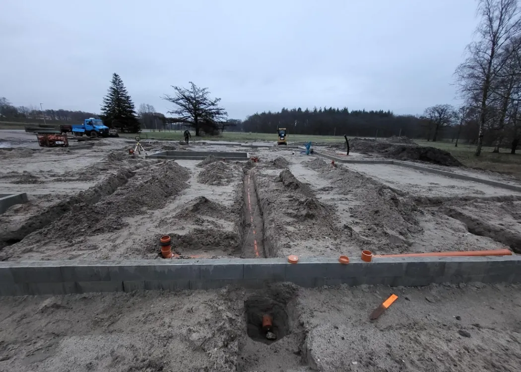 Baugrundstück der Tierarztpraxis Hohne im Harzklint. Erdreich mit sichtbaren Umrissen der Baufläche, Baugeräte und Markierungen im Vordergrund. Der Hintergrund zeigt eine Baumreihe bei grauem Himmel.