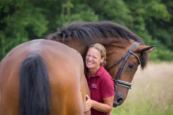 Eine Person mit ein Pferd von hinten. Die Person trägt ein rotes Poloshirt und umarmt das Pferd an der Brust. Die Frau lacht fröhlich in die Kamera.