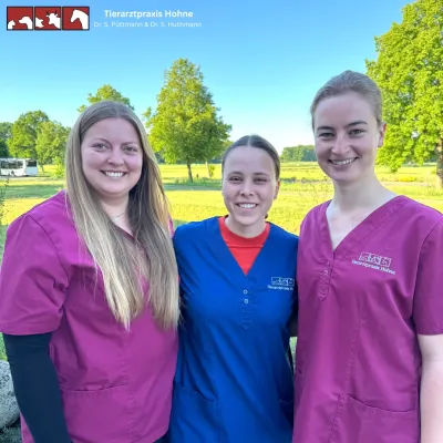 Drei Personen stehen nebeneinander im Freien, zwei tragen magenta und eine trägt ein blaues Oberteil. Alle lächeln fröhlich. Im Hintergrund sind Bäume und eine Wiese sichtbar.