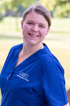 Lächelnde Frau mit zusammengebundenen blonden Haaren trägt ein blaues Oberteil mit dem Logo 'Tierarztpraxis Hohne'. Hintergrund zeigt unscharfe grüne Bäume.