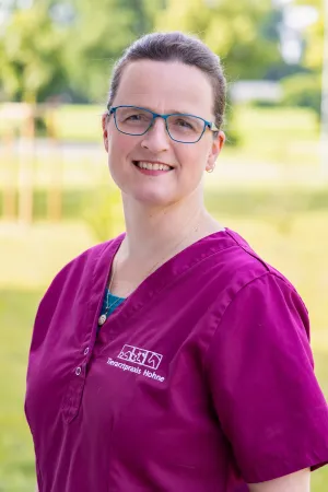 Lächelnde Frau mit braunen Haaren in magenta Arztkittel mit gesticktem Logo 'Tierarztpraxis Hohne'. Sie steht im Freien, der Hintergrund zeigt unscharfe grüne Bäume.