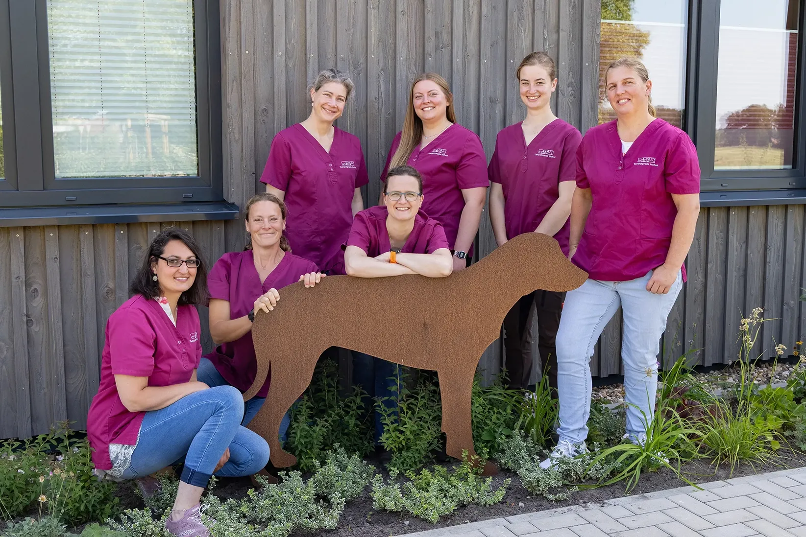 Sechs Personen in magenta Arztkitteln stehen und sitzen um eine rostfarbene Hundeskulptur. Sie befinden sich vor einer Holzfassade und stehen bzw. sitzen auf einer bepflanzten Fläche. Sie lächeln alle in die Kamera.