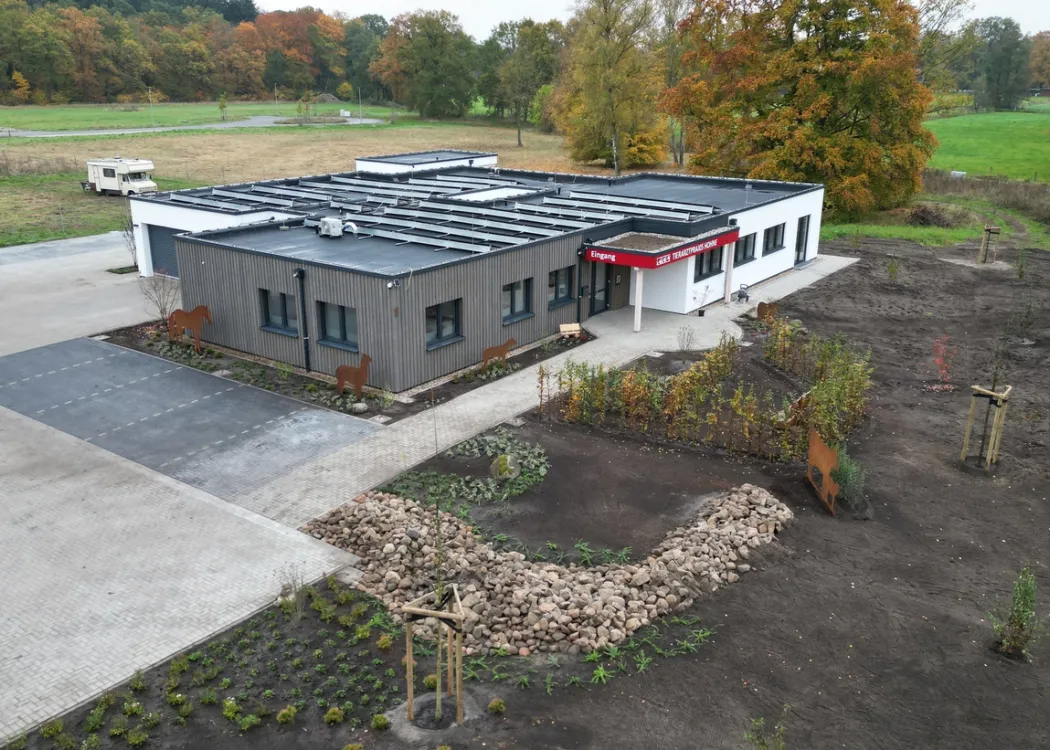 Luftaufnahme der Tierarztpraxis Hohne im Harzklint. Das Gebäude ist modern, flach und hellgrau, mit dunklem Dach, Solaranlage und rotem Vordach. Umgeben von Grünflächen und Wald im Hintergrund.