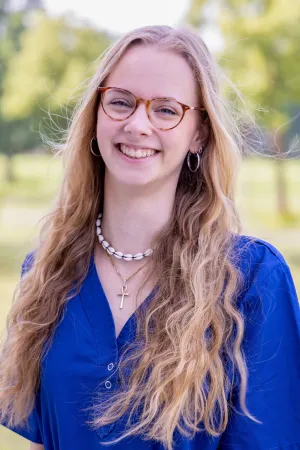 Frau mit langen, gewellten blonden Haaren trägt ein blaues Oberteil und eine Halskette mit Muscheln und einem Kreuzanhänger. Sie steht im Freien und lächelt.