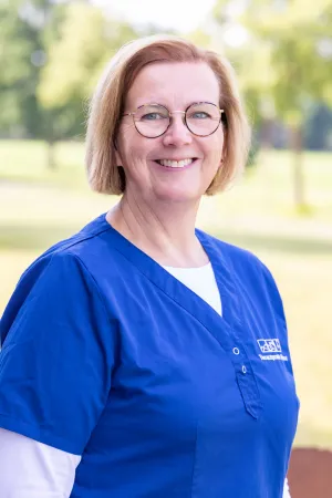 Frau mit blonden Haaren trägt ein blaues Arztkittel. Sie steht im Freien, im Hintergrund sind Bäume und Wiese sichtbar.