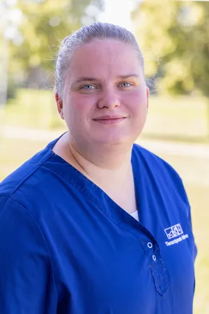 Leicht lächelnde Frau mit zusammengebundenen blonden Haaren trägt ein blaues Oberteil mit dem Logo 'Tierarztpraxis Hohne'. Hintergrund zeigt unscharfe grüne Bäume.