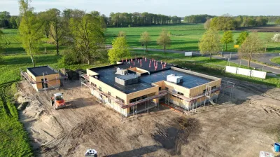 Bau einer Tierarztpraxis mit flachem Dach, Holzfassade und Baustellenzäunen. Umgeben von Wiese und Bäumen, mit Baumaschinen im Vordergrund.